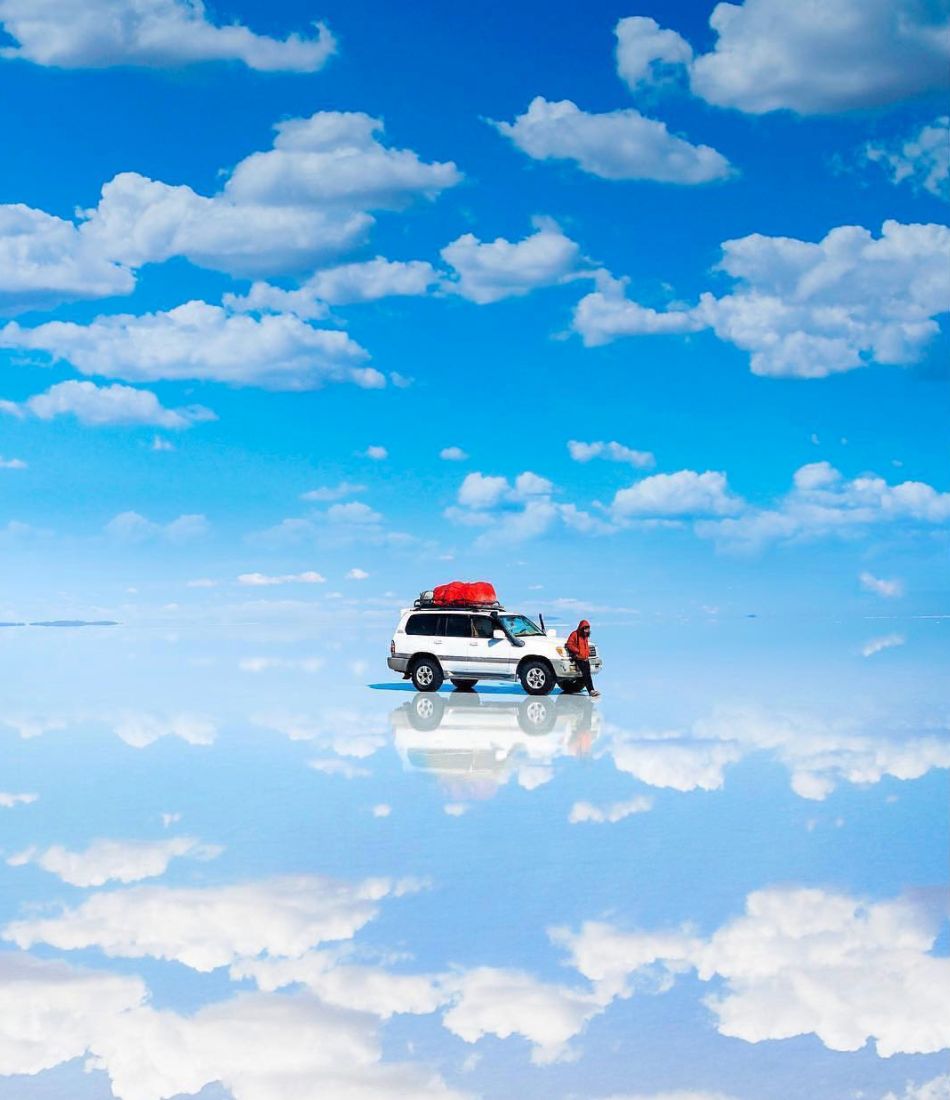 A tour jeep and a traveler reflected in the flooded Salar de Uyuni under a sky full of clouds, Bolivia
